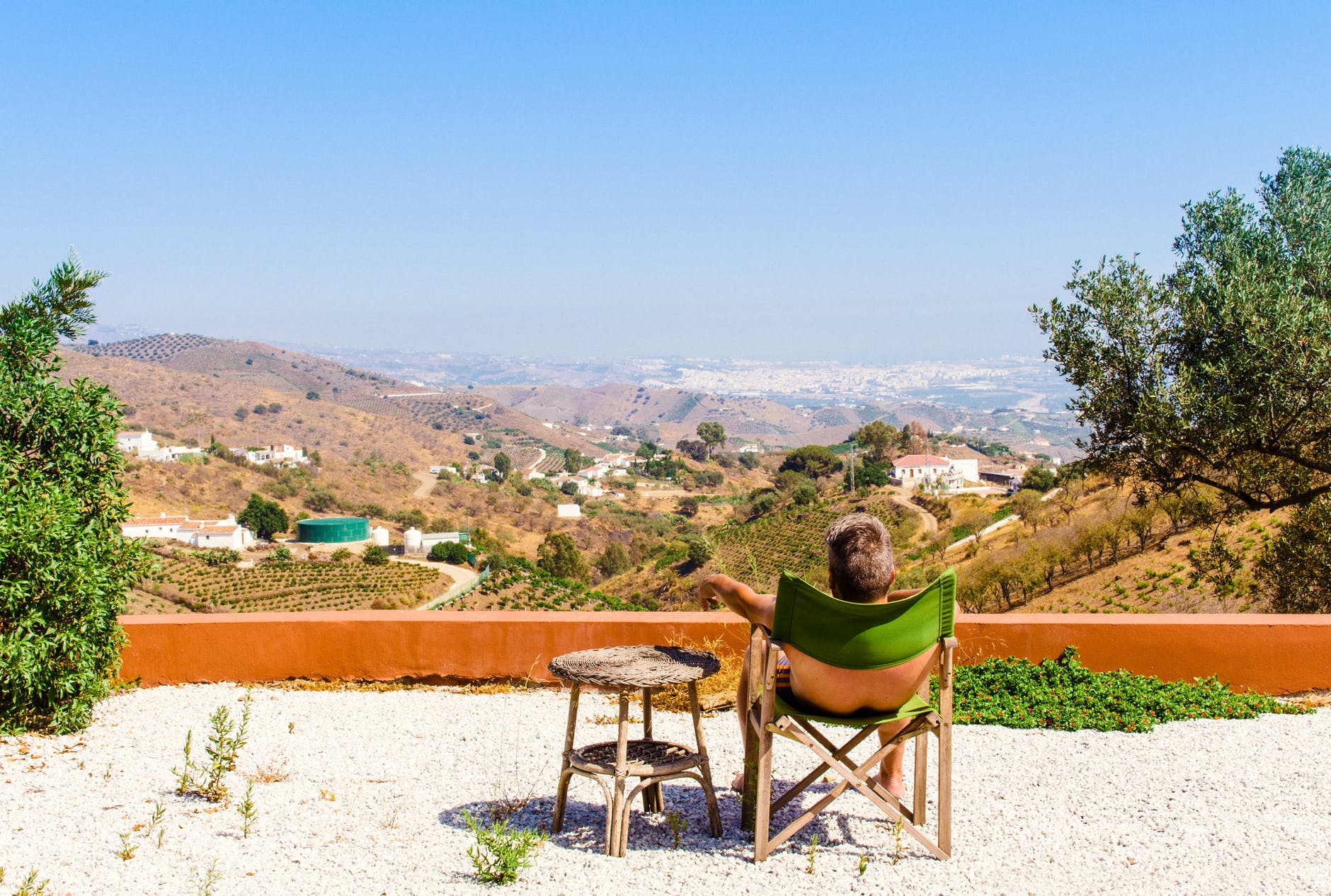 man sitting on green chair near trees and mountain under blue sky at daytime